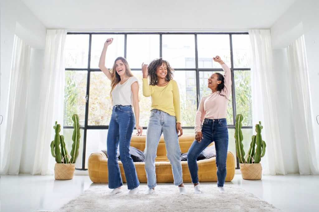 Three funny female friends dancing and singing on the sofa at home